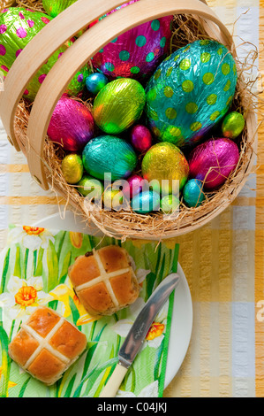 wooden basket filled with colorful foil wrapped chocolate easter eggs on yellow and white tablecloth Hot cross buns on plate Stock Photo