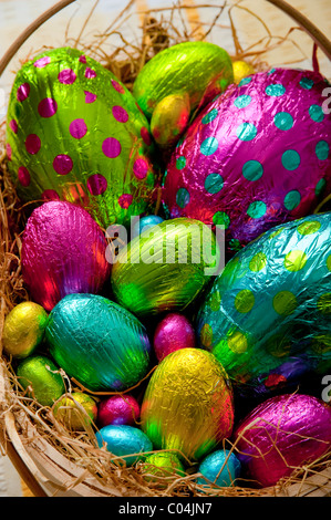 Traditional wooden basket filled with colorful foil wrapped chocolate easter eggs on yellow and white tablecloth Stock Photo
