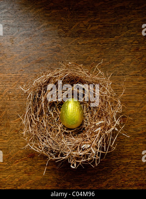 Single Gold foil wrapped chocolate easter egg in a hay birds nest on an oak table. Stock Photo