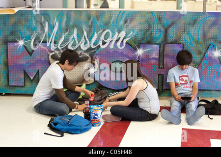 Hispanic boy and girl eat snacks while another boy sends text message on his cell phone during high school lunch break Stock Photo