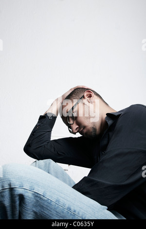 Young man wearing glasses sitting on the floor and holding his hand on his head Stock Photo
