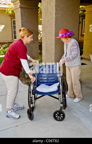 A uniformed Hispanic orderly holds a wheelchair for a red hatted Asian woman resident at a retirement home in Mission Viejo, CA Stock Photo