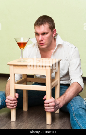 Drunken man sitting on floor with glass of alcohol and holding stool Stock Photo