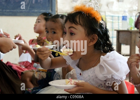 A 6 year old girl wearing a crown is eating a piece of birthday cake with a plastic spoon in a kindergarten class in Cambodia. Stock Photo