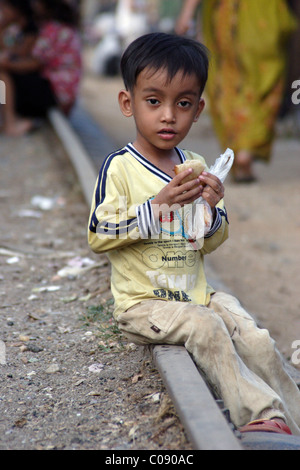 A young Asian boy is eating a sandwich while sitting on an abandoned stretch of railroad track in Phnom Penh, Cambodia. Stock Photo
