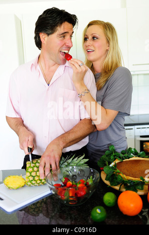 Man cutting a pineapple, young woman leaning on his shoulder and letting him bite into a strawberry Stock Photo