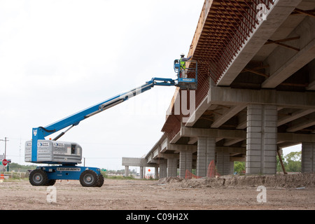 Construction worker wearing hard hat and safety vest stands in basket of boom lift to work on deck of highway bridge in Texas Stock Photo