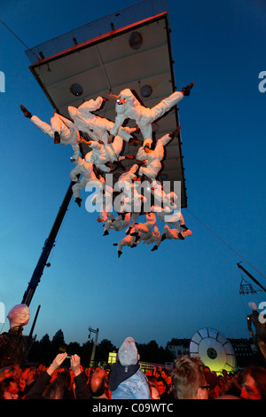 White-clad trapeze artists hanging upside down from a platform in the air, Global Rheingold, open-air theater by La Fura dels Stock Photo