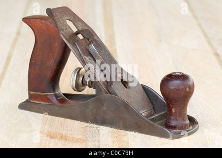 A vintage hand plane with a dark metal body and wooden handles rests on a light-colored wooden surface. The plane features a curved wooden front handle with a brass inlay and a rounded wooden knob at Stock Photo
