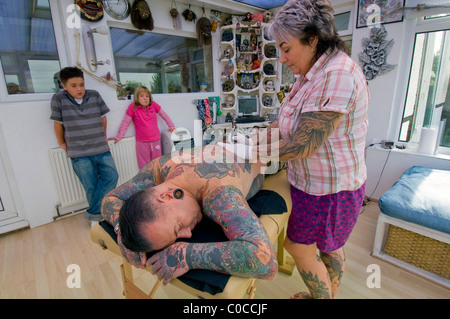 A woman tattooist works on a design on her husband's back, watched by their two bored children. Stock Photo