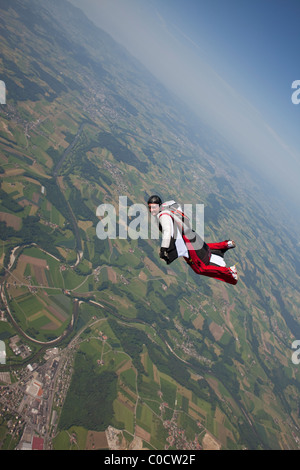 Skydiver within special wingsuit is flying over green fields in the blue sky. Stock Photo