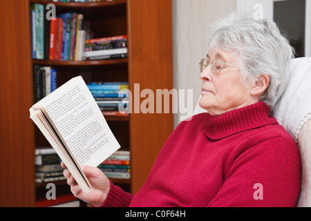 Someone elderly woman wearing a red jumper relaxing sat reading a paperback book in a living room with a bookcase behind. UK Stock Photo