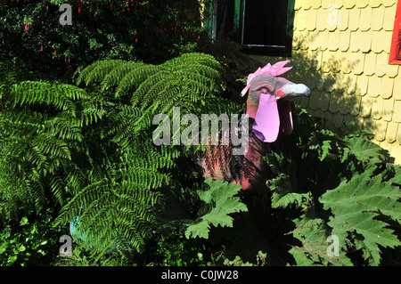 Sunny garden view of painted Basilisco in green gunnera and fern leaves in front of yellow shingles house Caulin, Chiloe, Chile Stock Photo