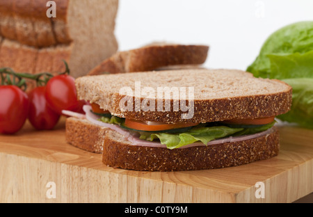 Ham and salad wholemeal sandwich freshly made on a cutting board with a loaf of bread, tomatoes and lettuce in the background. Stock Photo