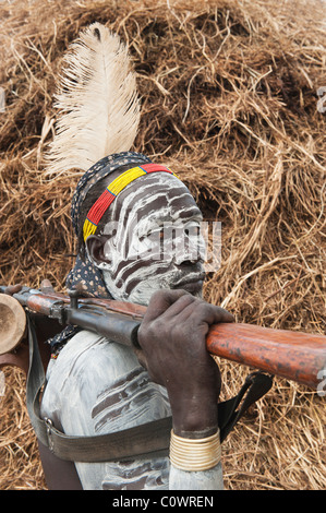 Karo warrior with body and facial paintings and a rifle in front of a hut, Omo river valley, Southern Ethiopia Stock Photo