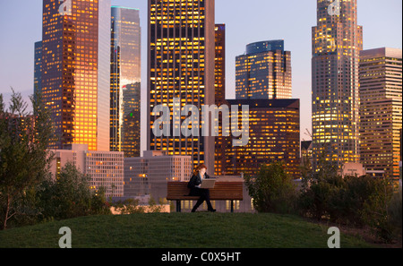 Female sitting on park bench working on laptop computer with Los Angeles city skyline in background at dusk, sunset Stock Photo