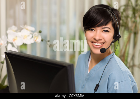 Smiling Attractive Multi-ethnic Young Woman Wearing Headset and Scrubs Near Her Computer Monitor. Stock Photo