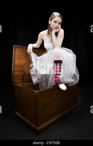 A teenage dancer wearing a white ballerina's dress and red striped socks sits on a wood trunk.