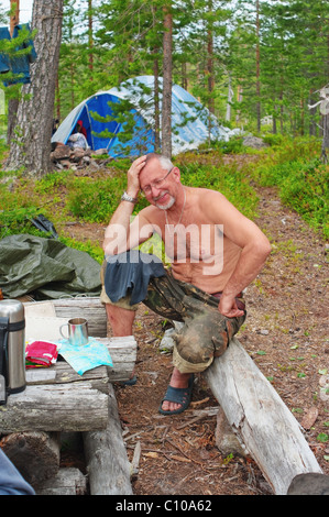 The smiling tourist sits on bench from logs in taiga wood. Stock Photo