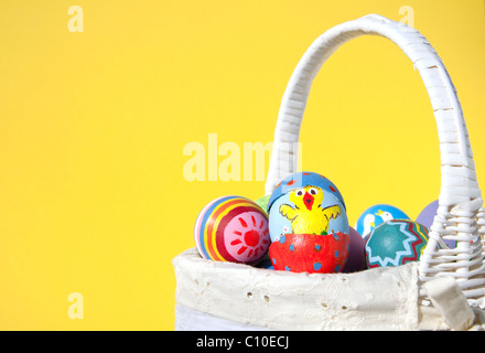 Easter chick painted on an egg shell peeking out of another painted egg shell, in a wicker basket with yellow background Stock Photo