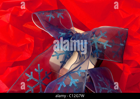 Vibrant holiday image of white jewelry box wrapped in a blue, snowflake accented ribbon against a background of red tissue paper Stock Photo