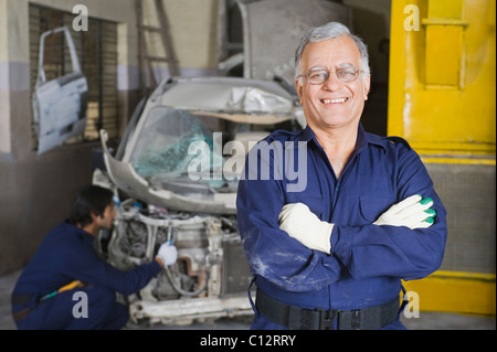 Portrait of an auto mechanic smiling with an apprentice repairing a car in the background Stock Photo