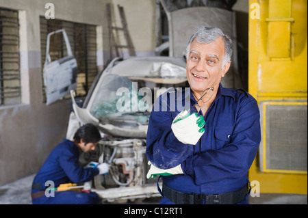 Portrait of an auto mechanic smiling with an apprentice repairing a car in the background Stock Photo