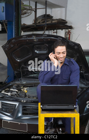 Auto mechanic talking on a mobile phone while using a laptop in a garage Stock Photo