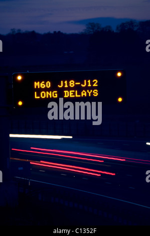 headlight trails of traffic on the A1/M motorway passing gantry warning sign of delays on the M60 ahead at night near leeds uk Stock Photo