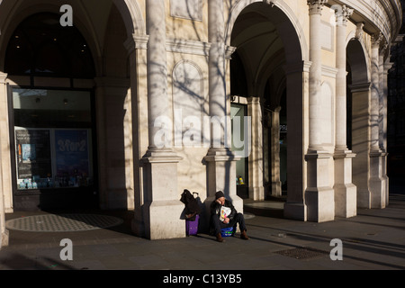 A sleeping Big Issue magazine seller sits alone without passing business in London's Trafalgar Square. Stock Photo