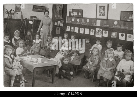 Original WW1 era postcard of Infant and nursery school children in classroom with their teacher, having a snack, or playing with toy teacups, circa 1913, 1914 1915 U.K. Stock Photo