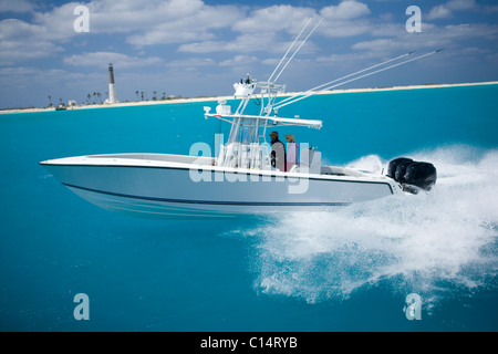 A fishing boat speeds through the blue water with the shore-line and lighthouse in the distance. Stock Photo