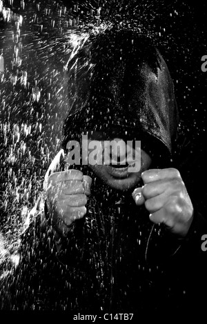 A young man wearing a hooded jacket poses in a wushu stance while it rains down on him. Stock Photo