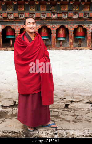 Young Buddhist monk in a maroon robe stands in front of a series of prayer wheels at his temple in the Paro Valley of Bhutan. Stock Photo