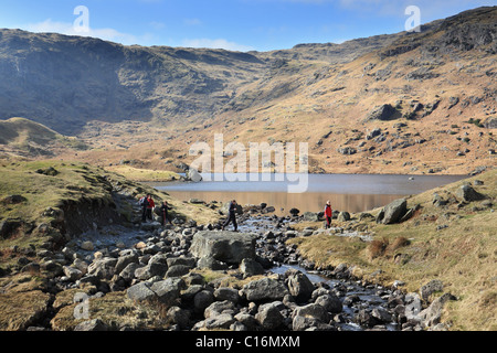 Fell walkers crossing stepping stones over Sourmilk Gill, Easedale Tarn in the background.  Near Grasmere English Lake District. Stock Photo