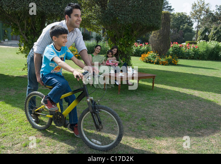 Man teaching his son to ride bicycle with his family sitting in the background Stock Photo