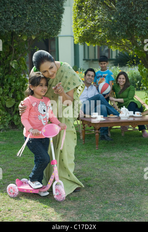 Woman teaching her granddaughter how to ride a push scooter with family in the background Stock Photo
