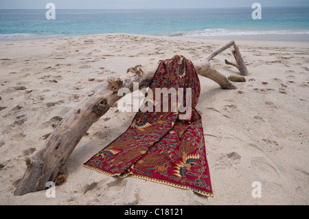 A batik sarong draped over a driftwood log on the white sand beach of Bias Tegal in Padang Bai, Bali Stock Photo