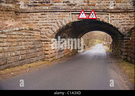 Old stone railway bridge with low warning signs over a country lane Stock Photo