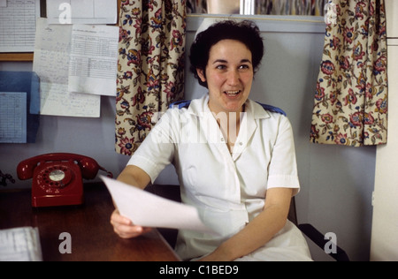 A young NHS nurse smiling in a white uniform sitting at a desk next to a red telephone holding a letter in a hospital administration office in the 1970s 1980s in Bronglais hospital Aberystwyth Wales UK   KATHY DEWITT Stock Photo