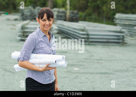 Young engineer woman supervising yard with rolled drawings in her hand Stock Photo