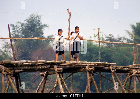 A group of 3 Laotian school girls are walking on a wooden bridge over a rural river in communist Laos. Stock Photo