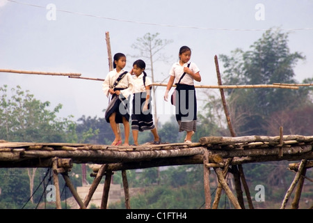 A group of 3 Laotian school girls are walking on a wooden bridge over a rural river in communist Laos. Stock Photo