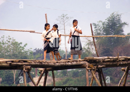 A group of 3 Laotian school girls are walking on a wooden bridge over a rural river in communist Laos. Stock Photo