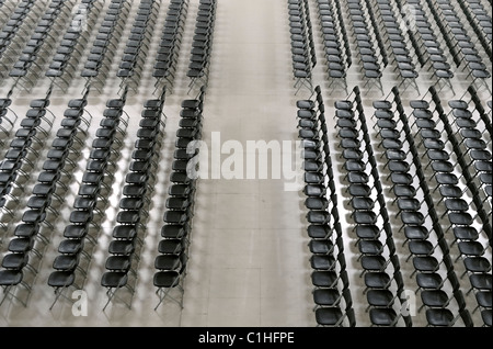 A high-angle black and white photograph showing row after row of empty black chairs neatly arranged in a large, open indoor venue. Stock Photo