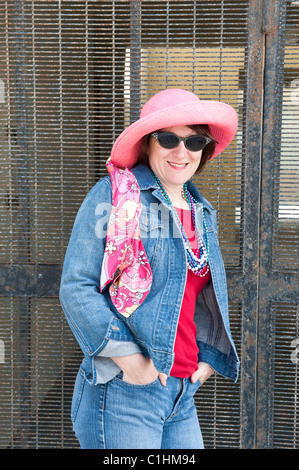 A woman poses against a metal security gate while wearing a colorful pink hat, vibrant scarf and sunglasses. Stock Photo