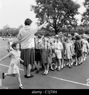 1960s. Female teacher holds a sign (the letter B) in her right hand  as she organises a group of young children to form a line in a school playground. Stock Photo