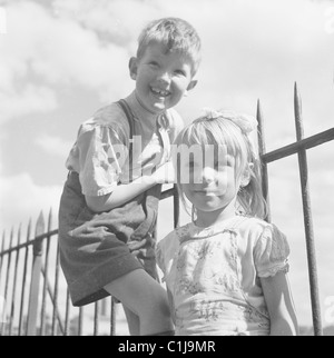 1950s, summertime, two children, a brother and sister by iron railings for a photo, the small boy in shorts climbing up, with a big smile on his face. Stock Photo