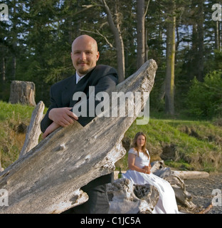 This smiling groom is leaning on some driftwood at a beach while his bride is sitting beautifully in the background. Stock Photo
