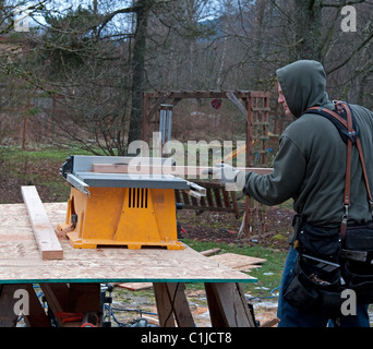 This hard working man is doing construction on site using power table saw. He is working during cold weather so wearing a hoodie Stock Photo